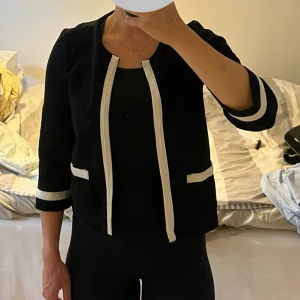 Svart kavaj/tröja  med vita detaljer från frk - Stilren svart kavaj från frk i storlek M med vita kontrastdetaljer längs öppningen, fickorna och ärmsluten. Modellen är rak och har trekvartsärm,. Materialet känns mjukt och har en lätt struktur