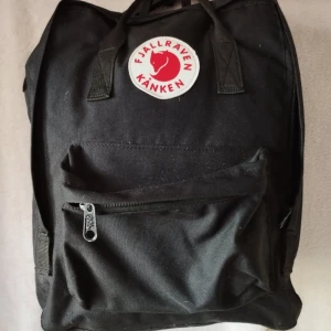 Svart Fjällräven Kånken ryggsäck - Klassisk svart Fjällräven Kånken ryggsäck med ikonisk rund röd och vit logotyp framtill. Ryggsäcken har ett stort huvudfack, en mindre ficka med dragkedja på framsidan och två handtag upptill. Tillverkad i slitstark polyester, perfekt för skola eller häng. 