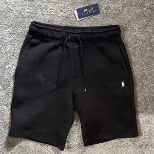 Svarta shorts från Polo Ralph Lauren - Storlek M .Svarta shorts från Polo Ralph Lauren med klassisk vit logobrodyr på benet. Modellen har snörning i midjan och två sidofickor. Mjukt material som känns skönt mot huden, perfekt för chill eller träning. Snygg och enkel design som funkar till allt.