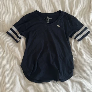 Mörkblå t-shirt Abercrombie Kids - Snygg mörkblå t-shirt från Abercrombie Kids med v-ringning och vita ränder på ärmarna. Klassisk logga broderad på bröstet. Skön passform och mjukt bomullsmaterial, perfekt för chill dagar. Sportig vibe med rundad nederkant.