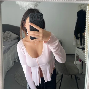 Ljusrosa långärmad topp med spets - Supersöt ljusrosa topp med lång ärm och croppad passform. Toppen har en djup rundringning, spetskant längs urringningen och framtill samt en liten rosett. Materialet är mjukt och lätt, med volangkant nertill.