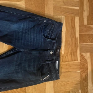 Mörkblå Replay Anbass jeans - Snygga mörkblå jeans från Replay, modell Anbass. Klassisk femficksdesign med Replay-logga på bakfickan och läderpatch i midjan. Jeansen har en smal passform och är tillverkade i stretchig denim för extra komfort.