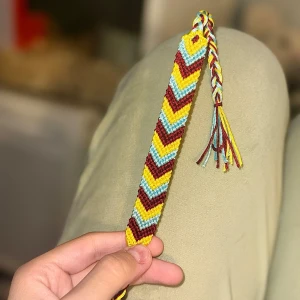 Handgjort vänskapsarmband  - Unikt handknutet vänskapsarmband med chevronmönster i vinrött, gult och ljusblått. Armbandet har flätade snören i matchande färger som avslutning.