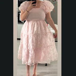 Ljusrosa klänning  - Superfin ljusrosa midiklänning med puffiga korta ärmar och smockad överdel. Kjolen är voluminös med lager och har ett romantiskt blommigt mönster i tyget. Perfekt för dig som gillar en feminin och drömmig stil.