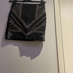 Svart glittrig kortkjol med paljetter - Supersnygg svart kortkjol med glittriga paljettdetaljer i geometriskt mönster framtill. Kjolen har en tight passform och dragkedja bak. Perfekt för dig som vill sticka ut med coola detaljer och glans på festen.