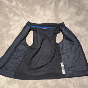 Ians väst - Svart fleeceväst från Patagonia med två dragkedjeförsedda fickor och blå detaljer på dragkedjorna. Västen har meshfoder på insidan och klassisk rak passform. Perfekt lager-på-lager-plagg för kyliga dagar.