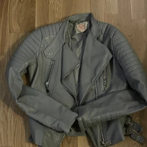 Grå skinnjacka med dragkedjor - Snygg grå skinnjacka från Royalty med coola detaljer som dragkedjor framtill, quiltade axlar och spännen i midjan. Jackan har en klassisk bikerstil med bred krage och långa ärmar. Perfekt för dig som vill ha en edgy look.