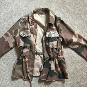 Camo parkajacka med paljettdetaljer XS - Snygg camouflage parkajacka från ONLY i bomull, med coola paljettdetaljer på bröstfickorna. Jackan har snörning i midjan, dragkedja och knappar framtill. Perfekt för dig som vill sticka ut med en unik och trendig look.