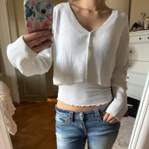 Vit croppad kofta Brandy Melville - Säljer en vit croppad kofta från Brandy Melville med v-ringning och knappar framtill. Tröjan är ribbad och har långa ärmar, perfekt för lager-på-lager. Enkel och stilren design som passar till jeans eller kjol. Skriv för fler bilder🩷