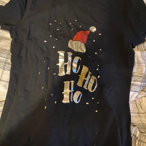Svart jul-tshirt från About You - Svart t-shirt med korta ärmar från About You, dekorerad med ett juligt tryck i guld och silver där det står 'Ho Ho Ho' och en röd tomteluva upptill. Materialet är mjuk bomull, perfekt för att maxa julkänslan med stil.