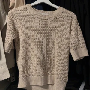 Beige stickad tröja från 157 - Säljer en beige stickad tröja från 157 med korta ärmar och snyggt hålmönster. Tröjan har rund hals och breda ribbade muddar vid ärmslut och nederkant. Perfekt för dig som gillar en chill och stilren look.