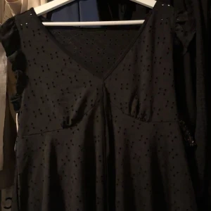 Svart kortklänning med volangärm - Säljer en svart kortklänning med v-ringad hals och små volanger på axlarna. Klänningen har ett diskret prickigt mönster och är tillverkad i ett mjukt bomullstyg. Perfekt för dig som gillar enkel men snygg stil med lite extra detaljer.