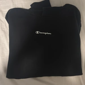 Champion hoodie - Svart hoodie från Champion med vit logga på bröstet och snygga band med Champion-tryck längs axlarna. Klassisk huva och stor magficka framtill. Perfekt för chill eller streetwear. Mjuk och skön i bomullsmix, normal passform och långärmad.