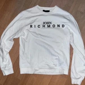 Vit sweatshirt - Säljer en stilren vit sweatshirt från John Richmond med svart logotyptryck på bröstet. Tröjan har rund hals, långa ärmar och ribbade muddar vid ärmslut och nederkant. Perfekt för dig som gillar cleana och enkla designer.