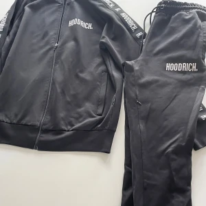 Svart tracksuit från Hoodrich - Svart tracksuit från Hoodrich med vit logga på både jacka och byxor. Jackan har dragkedja och ribbade muddar, byxorna har resår och snörning i midjan. Snygga loggoband längs ärmar och ben ger en sportig vibe. Perfekt för dig som gillar streetwear.