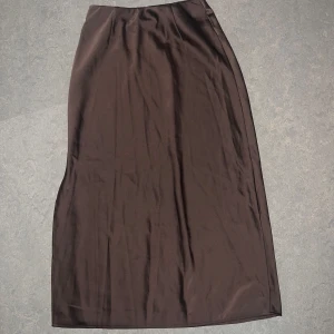 Brun satin lång/medel kjol från bikbok - Snygg brun långkjol i satin med slät och glansig yta. Kjolen har en rak passform och faller mjukt längs benen. Perfekt för dig som gillar en stilren och enkel look. Öppen vid sidan 