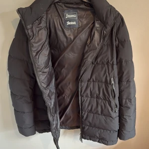 Svart pufferjacka från Herno  - Snygg svart pufferjacka från Herno med huva och dragkedja. Jackan har quiltad design och två fickor med dragkedja framtill. Perfekt för kalla dagar när du vill hålla stilen och värmen. Klassisk passform och stilren look.