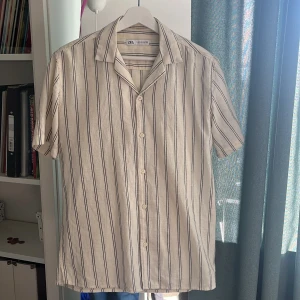 Randig kortärmad skjorta från Zara - Snygg kortärmad skjorta från Zara i beige med svarta vertikala ränder. Skjortan har klassisk krage, knäppning framtill och är tillverkad i ett luftigt linnematerial. Perfekt för varma dagar när du vill ha en avslappnad men stilren look.