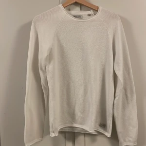 Vit stickad tröja från Jack & Jones - Vit långärmad tröja från Jack & Jones med rund hals och stickad struktur. Tröjan har en enkel och clean design, perfekt för dig som gillar minimalistisk stil. Materialet är mjukt och andas, vilket gör den skön att bära under kyliga dagar. 