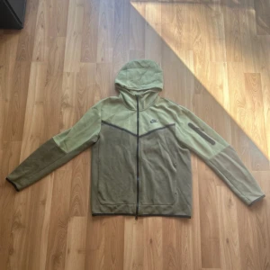 Nike hoodie i olivgrönt med dragkedja - Nike hoodie i två nyanser av olivgrönt med svart logga på bröstet. Tröjan har hel dragkedja, huva och en snygg detalj med dragkedjeficka på ena ärmen. Materialet är mjuk bomull och polyester, perfekt för chill eller träning.