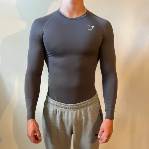 Mörkgrå Gymshark comp shirt  - Slimmad och mörkgrå långärmad träningströja från Gymshark i storlek S. Tröjan har en stilren design med diskret logga på bröstet och är gjord i stretchigt syntetmaterial som sitter tight mot kroppen. Perfekt för gymmet eller sport.  Jag är 191 och väger 91 kg och den är lite väl tight på mig