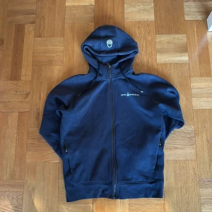 Marinblå hoodie från Sail Racing - Säljer en marinblå hoodie från Sail Racing med dragkedja och huva med snörning. Tröjan har två fickor med dragkedja och logga på bröstet. Perfekt för chill dagar eller när du vill ha en sportig look.