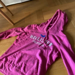 Rosa hoodie från Hollister XS - Säljer en snygg rosa hoodie från Hollister i storlek XS. Tröjan har en stor magficka, huva med snörning och vit broderad logga samt en blå fågel på bröstet. Perfekt för chill dagar och har en skön, mjuk känsla tack vare bomullsmaterialet.