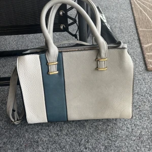 Beige och blå axelväska med gulddetaljer - Snygg axelväska i beige skinnimitation med en mörkblå rand på framsidan och guldfärgade metalldetaljer. Väskan har både handtag och justerbar axelrem, flera fack med dragkedja och ljus insida med logga. Perfekt för dig som vill ha stilren och praktisk väska.