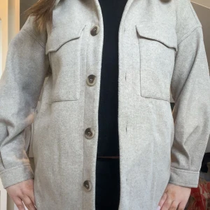 Beige overshirt med stora fickor - Snygg beige overshirt med stora bröstfickor och knappar framtill. Jackan har en rak passform och är tillverkad i ett mjukt ullblandat material. Perfekt att slänga över en hoodie eller t-shirt för en chill look.