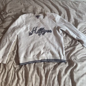 Vit Hilfiger sweatshirt med brodyr - Vit sweatshirt från Tommy Hilfiger med broderad Hilfiger-logga i mörkblått på bröstet. Tröjan har långa ärmar och ribbade muddar med marinblå ränder vid nederkant och ärmslut. Tillverkad i mjuk bomull, perfekt för en chill och stilren look.