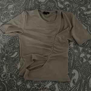 Beige glittrig topp med rynkdetalj - Säljer en beige topp med glittrigt tyg och snygga rynkdetaljer på framsidan. Toppen har korta ärmar och en rund halsringning. Perfekt för dig som vill sticka ut lite extra med en stilren men ändå unik look.