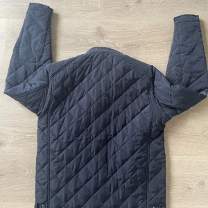 Quiltad blå jacka från Replay - Snygg quiltad jacka från Replay i mörkblått med dragkedjor på ärmar och fickor. Jackan har ett rutigt mönster, hög krage med knapp och en bröstficka med dragkedja. Perfekt för dig som gillar stilren och sportig look.(PRIS KAN DIKSUTERAS) storlek m