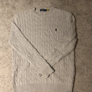 Ljusgrå kabelstickad tröja Polo Ralph Lauren Storlek L - Kabelstickad ljusgrå tröja från Polo Ralph Lauren med klassisk rund halsringning och broderad logga på bröstet. Tröjan har ribbade muddar och är långärmad, perfekt för lager-på-lager under kyliga dagar.