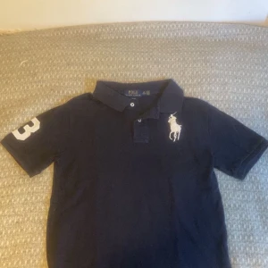 Marinblå Polo Ralph Lauren piké  - Snygg marinblå piké från Polo Ralph Lauren med klassisk krage och två knappar. Stor vit broderad logga på bröstet och en vit siffra 3 på ärmen. Classic fit och tillverkad i mjuk bomull. Perfekt för dig som gillar stilrena och sportiga plagg. Hör av er vid frågor och funderingar. Kan förhandla om pris vid snabb affär 