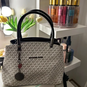Michael Kors handväska med MK-mönster - Snygg handväska från Michael Kors i beige och grått med ikoniskt MK-mönster över hela väskan. Väskan har svarta handtag, dragkedja upptill och en dekorativ MK-nyckelring. Perfekt storlek för att få plats med allt du behöver.