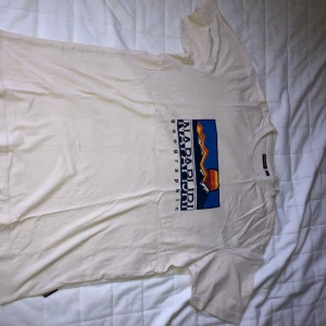 Vit Napapijri t-shirt med tryck - Vit t-shirt från Napapijri med stort färgglatt grafiskt tryck på bröstet. Klassisk rund halsringning och korta ärmar. Tillverkad i mjuk bomull, perfekt för en avslappnad och stilren look.