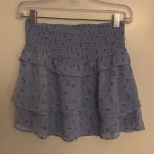 Blå blommig volangkjol Gina Tricot använd 1 gång - Supersöt blå kortkjol från Gina Tricot med små blommor och volanger. Kjolen har smock i midjan för en bekväm passform och är gjord i ett lätt, flowy material. Perfekt för dig som gillar en feminin och trendig stil.