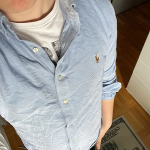 Blå skjorta från Ralph Lauren - Snygg ljusblå skjorta från Ralph Lauren i slim fit-modell. Klassisk oxfordväv i bomull med broderad polospelare på bröstet. Skjortan har långa ärmar, knappar framtill och en stilren, tidlös look som passar till jeans eller chinos.                            Osäker på pris så kom med förslag!!!
