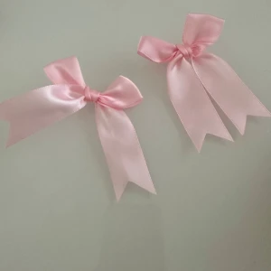 Rosa satinrosetter med clips - Två söta ljusrosa rosetter i satin med metallclips på baksidan. Perfekta att fästa i håret som diadem eller håraccessoar. Rosetterna har långa band och en glansig finish som ger en gullig och trendig look.