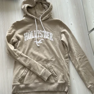Beige hoodie från Hollister med huva - Säljer en beige hoodie från Hollister med broderad logga framtill. Tröjan har huva med snörning, känguruficka och ribbade muddar. Perfekt för en chill och avslappnad stil. Mjuk insida och klassisk passform.