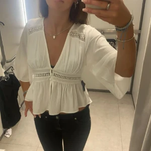 Vit blus med v-ringning från Zara - Superfin vit blus från Zara med djup v-ringning och knappar framtill. Blusen har vida trekvartsärmar, broderade detaljer och lätt rynkad midja som ger en snygg siluett. Perfekt att styla med jeans eller kjol för en trendig look.