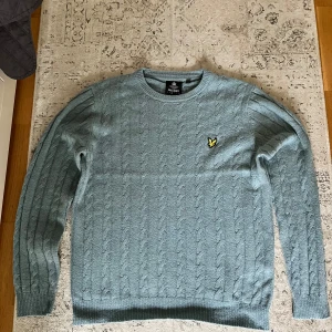 Lyle & Scott Stickad Tröja (NY) - Säljer en ljusblå stickad tröja från Lyle & Scott med klassiskt kabelstickat mönster och rund hals. Tröjan har lång ärm och den ikoniska gula fågel-loggan broderad på bröstet. Perfekt för dig som gillar stilren och tidlös design. Helt ny, aldrig använd, nypris: 1049 kronor. Köpt på Boozt för ett år sen.
