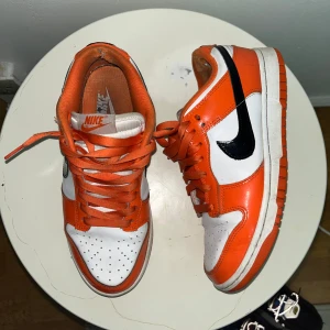 Nike Dunk Low orange och vita sneakers - Nike Dunk Low sneakers i orange och vitt med svart swoosh. Klassisk siluett med lågt skaft, perforerad tå och snörning. Ovandel i skinn och syntet, orange yttersula och matchande orange skosnören.