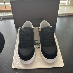 Burberry svarta sneakers med rutig detalj - Snygga svarta sneakers från Burberry med klassisk rutig panel i beige, svart och vit på sidan och hälen. Skorna har snörning, rund tå och vit platt sula. Materialmix av skinn och textil ger en lyxig känsla och cool kontrast. Perfekta för dig som gillar stilrena och trendiga skor.