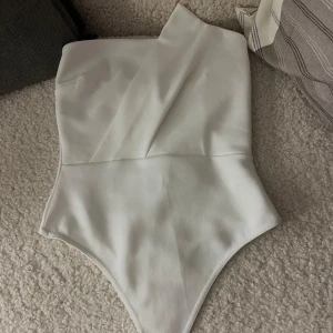 Vit body från Boohoo - Snygg vit body från Boohoo i storlek 36. Bodyn har en stilren omlott-design framtill och är axelbandslös för en clean look. Materialet känns mjukt och har lätt stretch, perfekt för att matcha med jeans eller kjol. Enkel att styla och superfräsch i sommar.