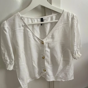 Vit blus med v-ringning från Divided - Vit blus från Divided med v-ringad hals och tre snygga knappar framtill. Blusen har korta ärmar med smock vid ärmslutet och är gjord i ett mjukt, lätt material som känns skönt mot huden. Perfekt för dig som gillar en clean och fräsch look.