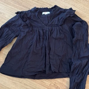 Svart blus med volang från Gina Tricot - Säljer en svart blus från Gina Tricot med volangdetaljer vid axlarna och v-ringad hals. Blusen har långa ärmar med spetsinslag och lätt rynkade partier som ger en cool och avslappnad vibe. Perfekt för dig som gillar stilrena plagg med lite extra detaljer.