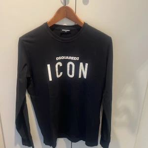 Svart långärmad tröja DSQUARED2 ICON - Svart långärmad tröjai från DSQUARED2 med stor vit 'ICON'-print på bröstet. Tröjan är tillverkad i 100% bomull och har en klassisk rund hals. Perfekt för dig som gillar streetstyle och vill sticka ut med ett tydligt statement.