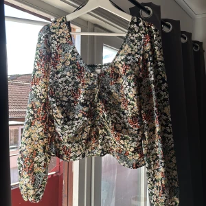 Blommig croppad blus med snörning - Superfin blommig blus med croppad passform och långa puffärmar. Toppen har snörning framtill och elastiska muddar vid ärmslut. Färgerna är svart, beige, röd och grön med ett detaljerat blommönster. Perfekt för dig som gillar romantisk och trendig stil.