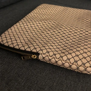 Brun/guldig datorväska med grafiskt mönster - Snygg brun/guldig datorväska med svart grafiskt mönster och guldfärgad dragkedja. Väskan är platt och rektangulär, perfekt för laptop eller dokument. Helt oanvänd och köpt från Zalando, passade nämligen ej min dator. Längd 35cm och bredd 25cm.